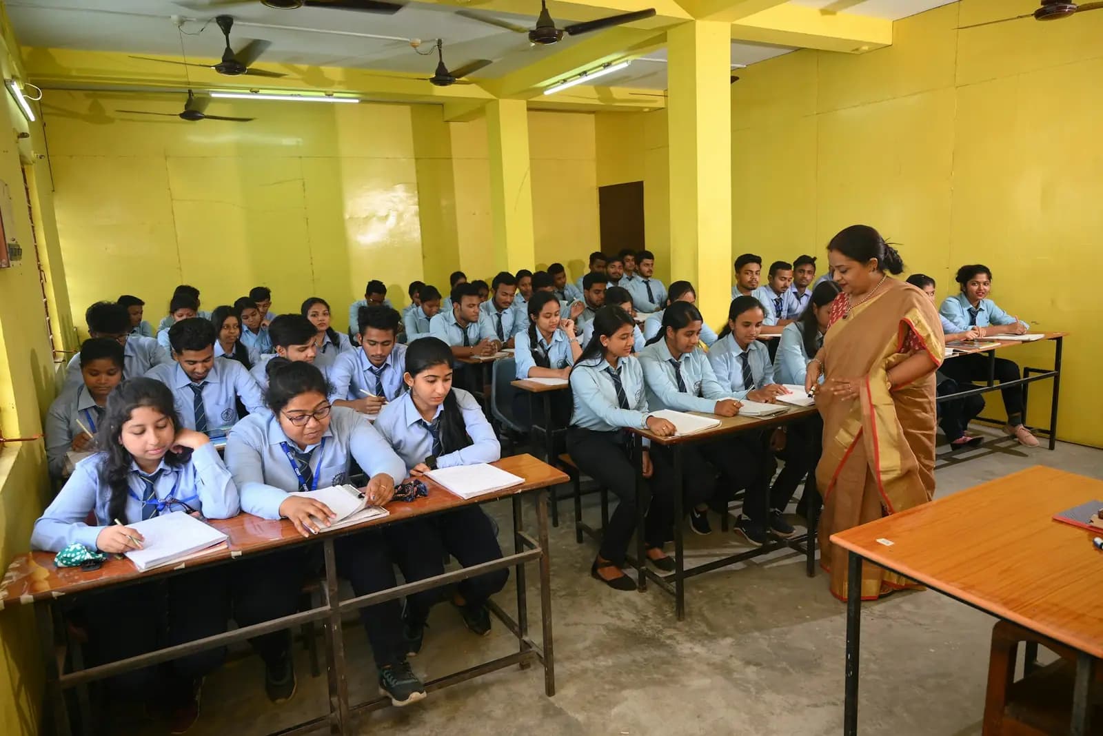 A wide view of a classroom where students in light blue uniforms write at their desks while a teacher in a sari walks between the rows