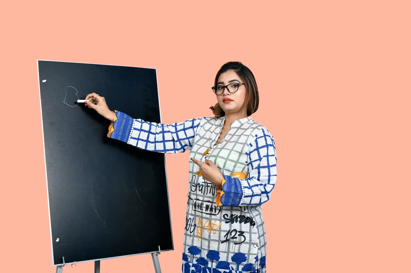 A woman in glasses and a checked kurta writes on a small blackboard on an easel and turns to smile
