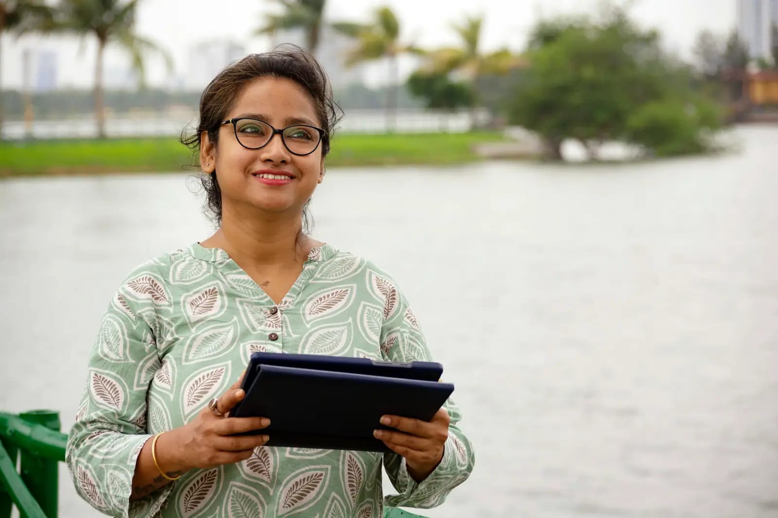 A young woman in glasses and a green kurta holds a tablet and smiles, with a lake and palm trees behind her