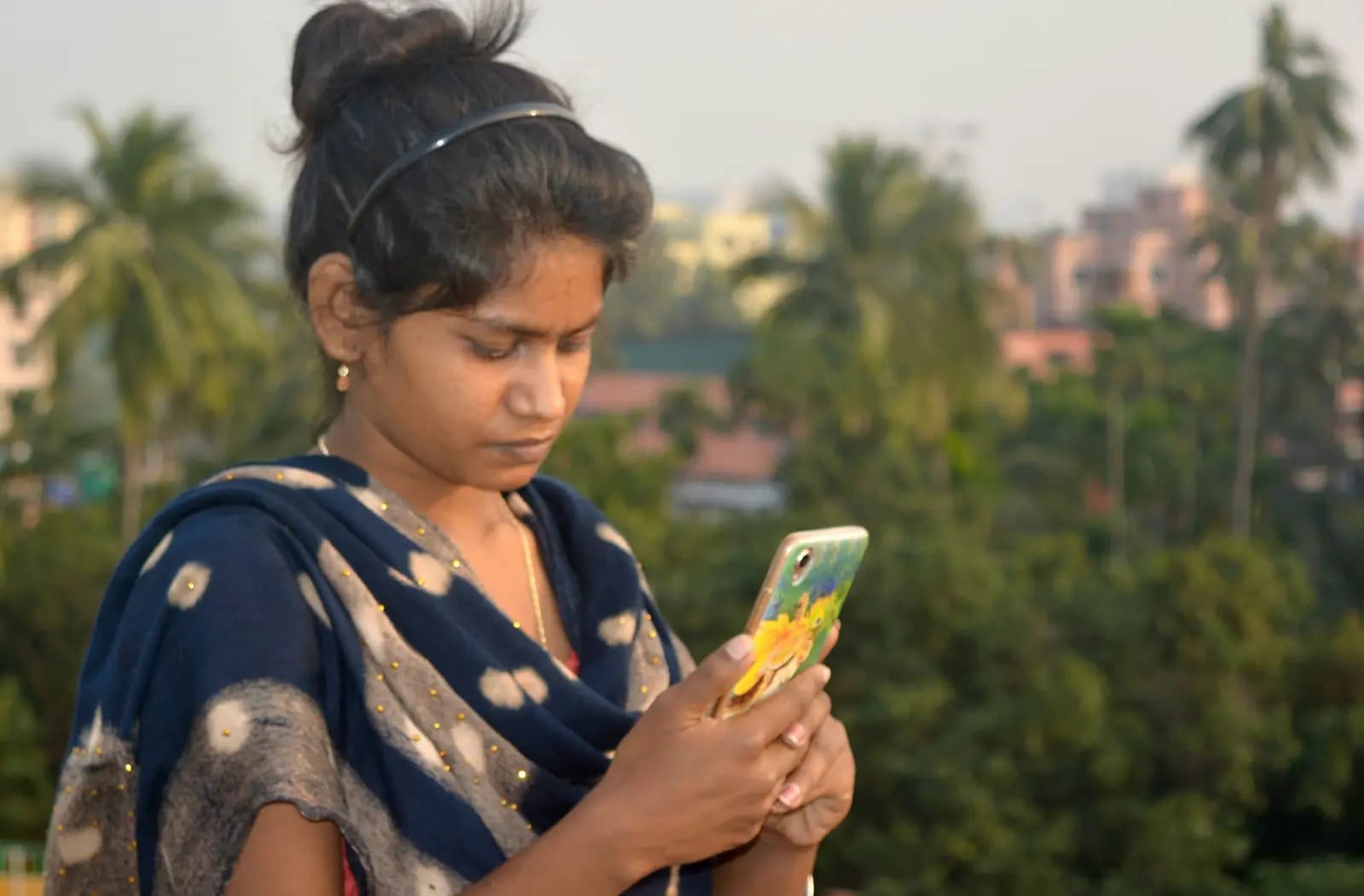 A young woman in a dupatta looks down at her phone as she types a message, trees and rooftops behind her