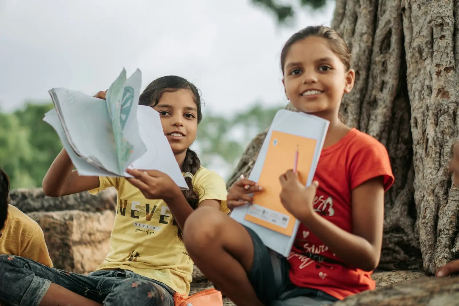 Two girls sit outdoors under a tree, one holding up an open notebook and the other smiling with her school folder